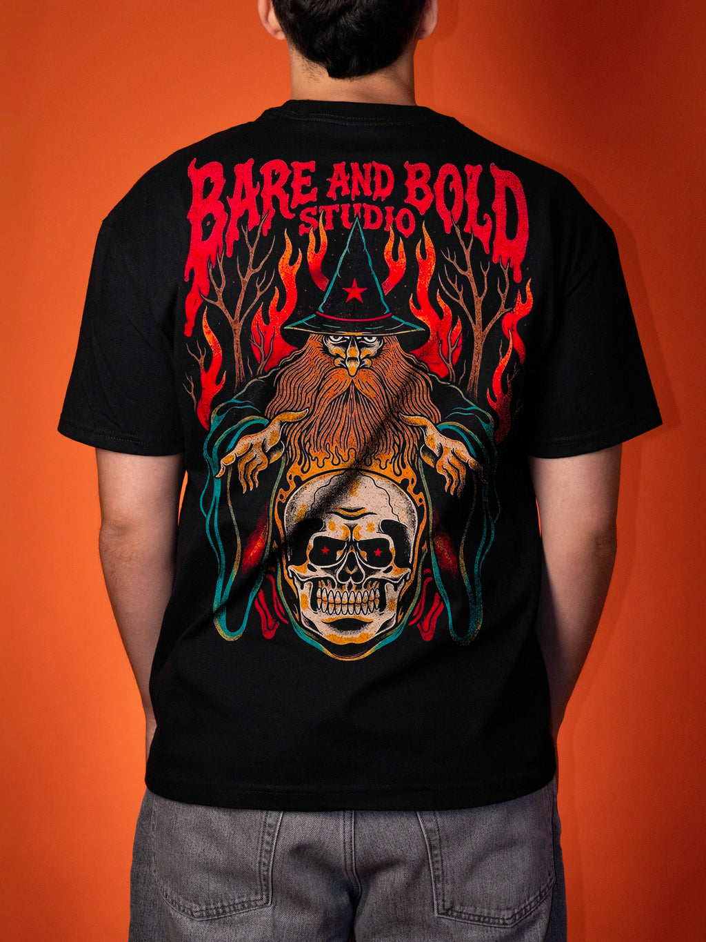 A person shown from the back wearing a black graphic t-shirt with a large red, orange, and yellow wizard and skull design, set against a solid orange background.