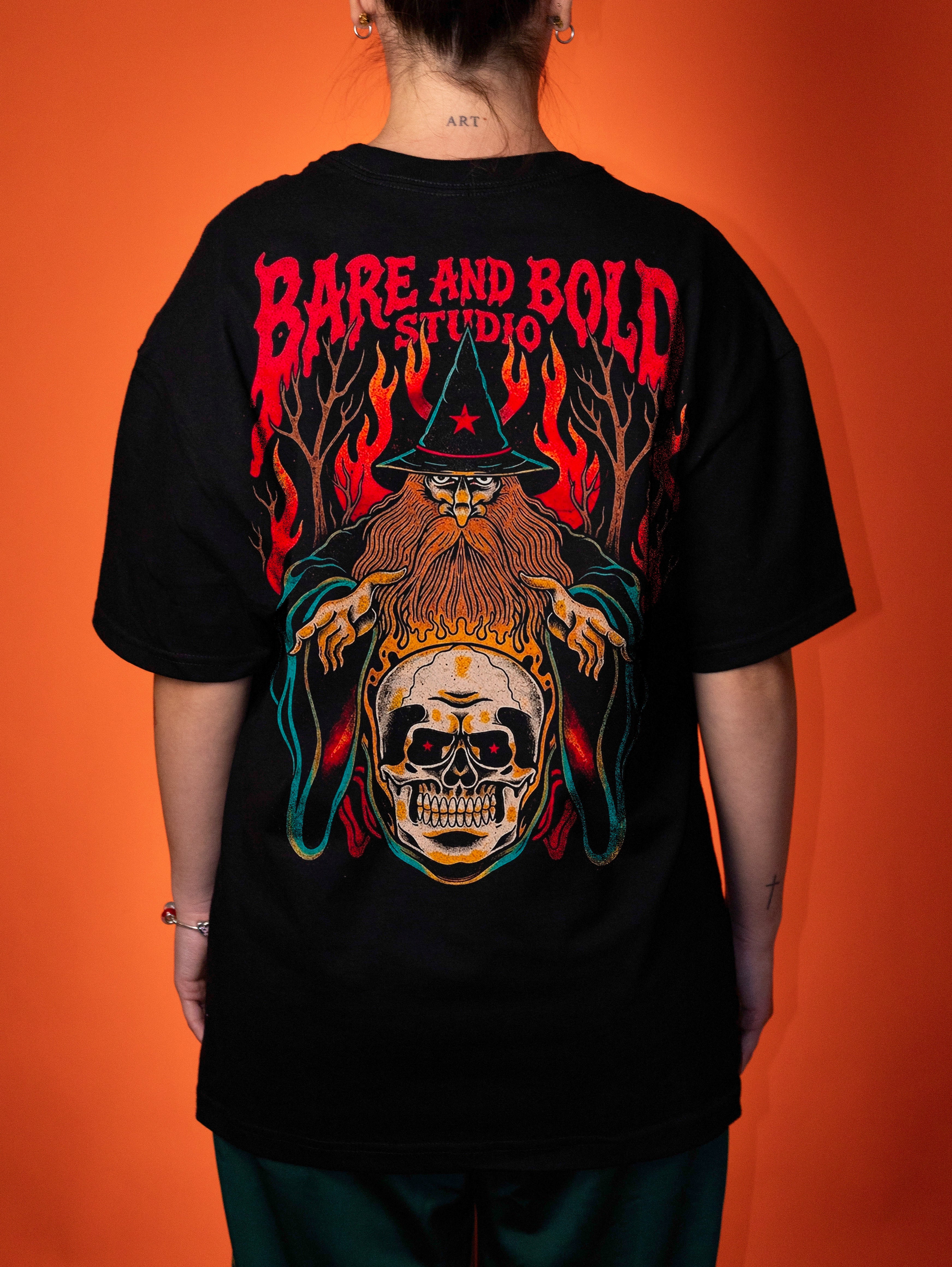 A person viewed from the back wearing a black graphic T-shirt with a large colorful design featuring the text 'BARE AND BOLD STUDIO' above a wizard, flames, and a skull. The background is a solid orange wall.