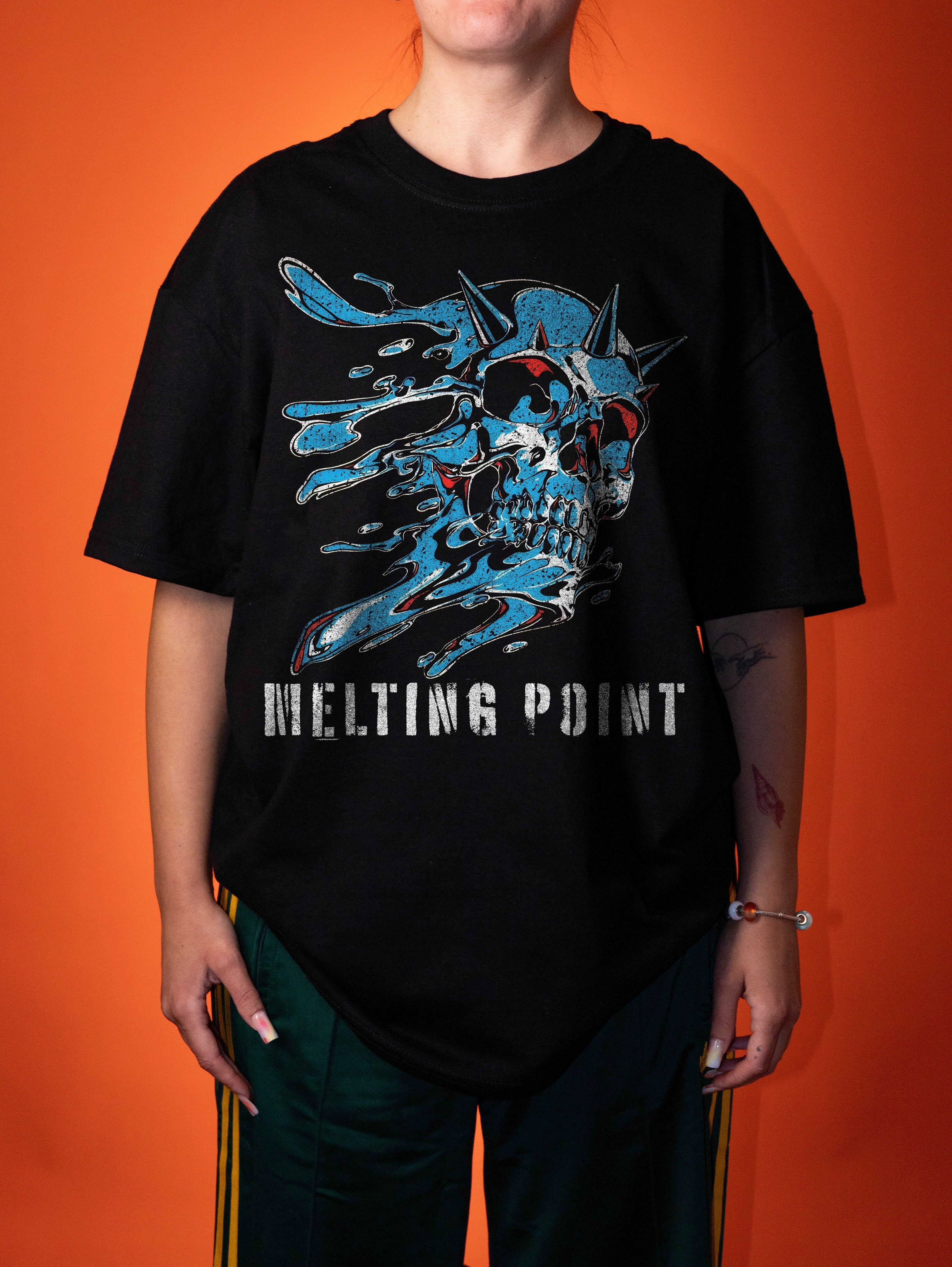 A person wearing an oversized black graphic T-shirt with a blue melting skull design and the words 'MELTING POINT' printed across the front, paired with dark green track pants, set against a solid orange background.