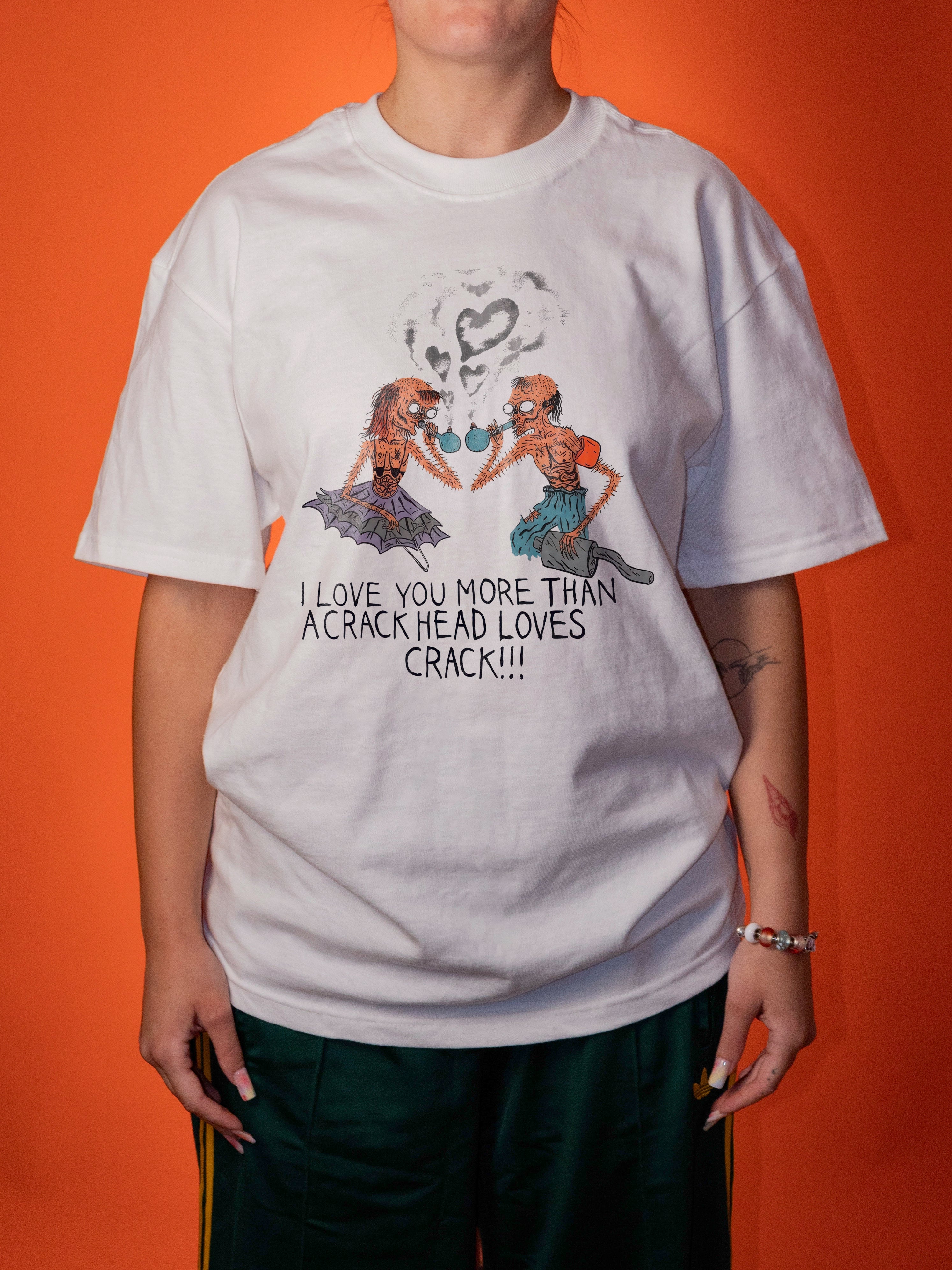 A person wearing an oversized white graphic T-shirt with a colorful illustration of two women and the text 'I LOVE YOU MORE THAN A CRACK HEAD LOVES CRACK!!!' against a solid orange background.