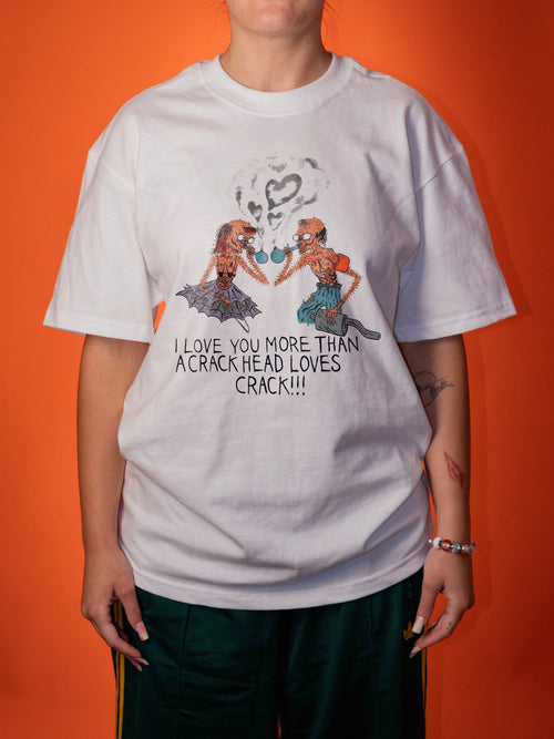 A person wearing an oversized white graphic T-shirt with a colorful illustration of two women and the text 'I LOVE YOU MORE THAN A CRACK HEAD LOVES CRACK!!!' against a solid orange background.