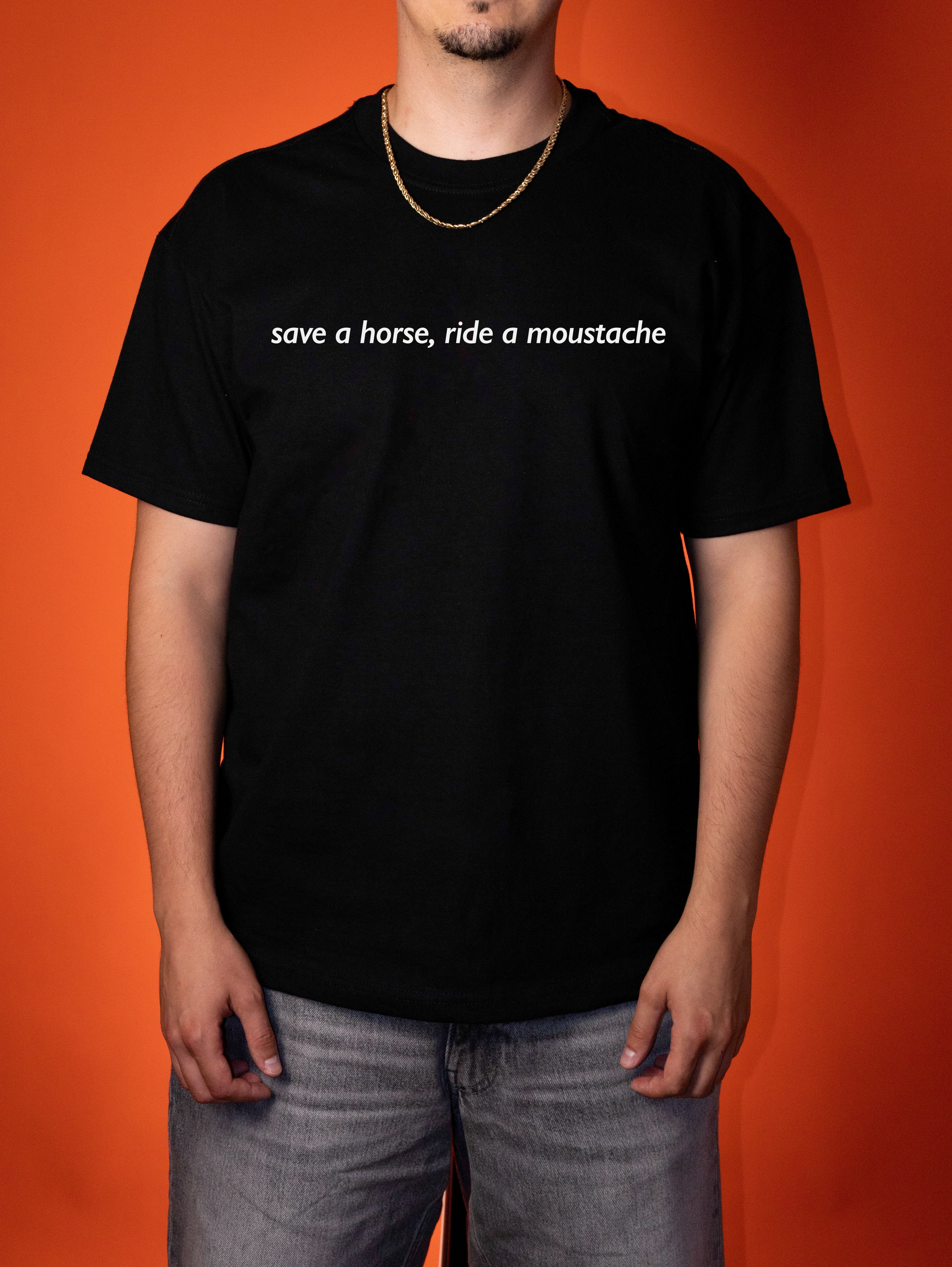A man wearing a black crewneck t-shirt with the white printed text 'save a horse, ride a moustache' on the chest, paired with gray jeans, set against a solid orange background.