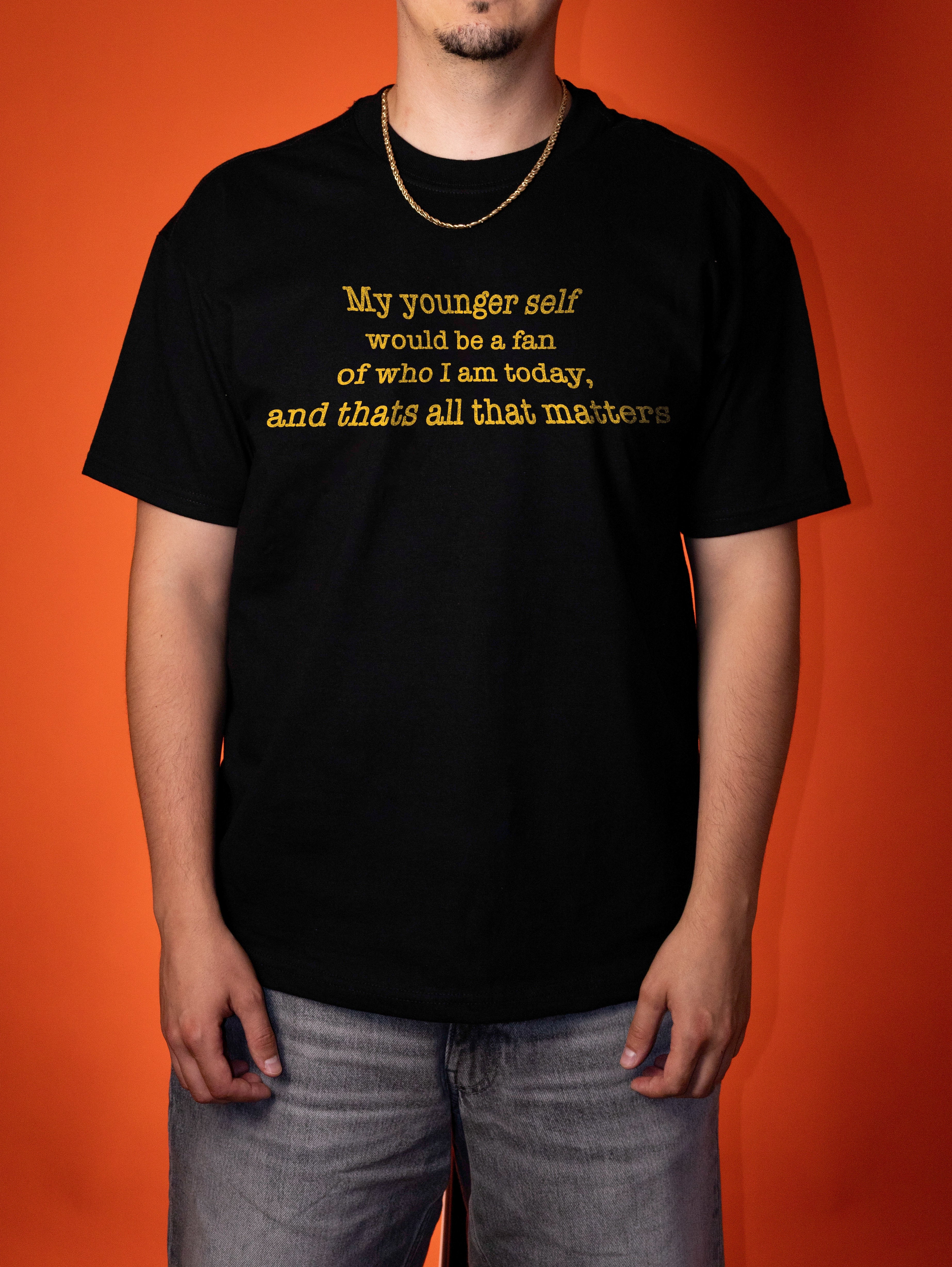 A man wearing a black short-sleeve t-shirt with yellow text that reads 'My younger self would be a fan of who I am today, and thats all that matters' against a solid orange background.