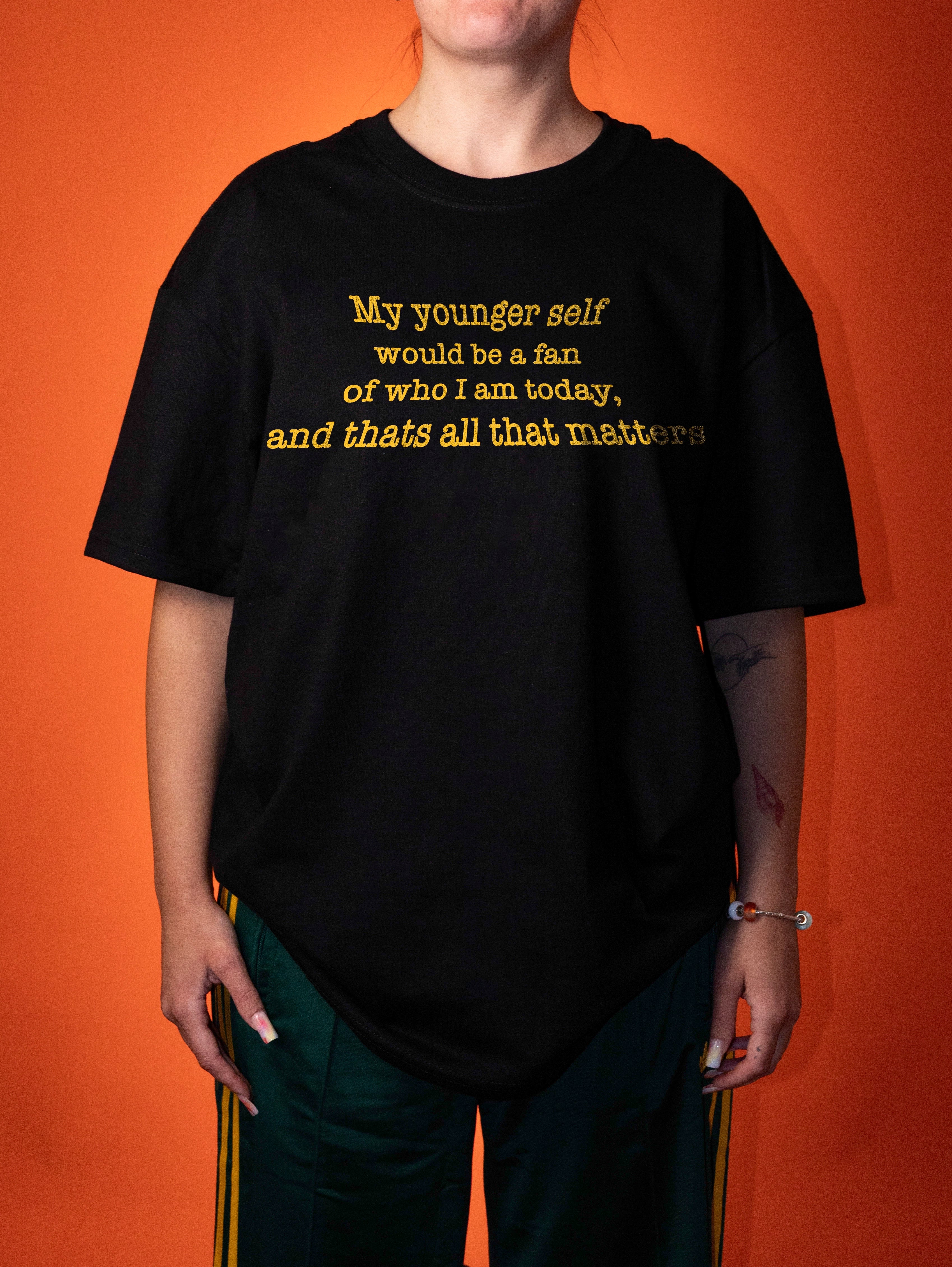A person wearing an oversized black graphic T-shirt with yellow text that reads, 'My younger self would be a fan of who I am today, and thats all that matters,' posed against a solid orange background.