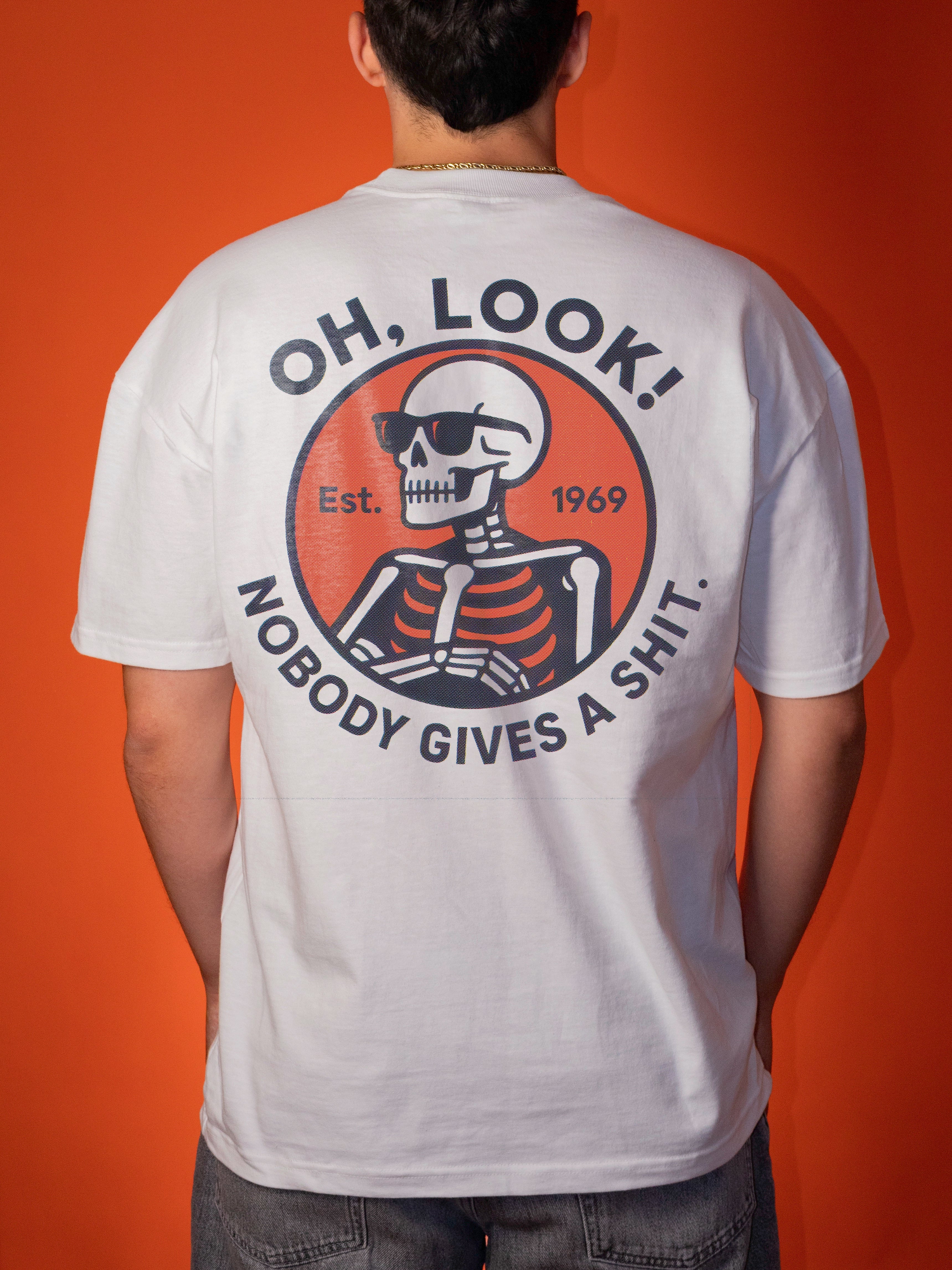 A person viewed from behind wearing a white graphic T-shirt with a large circular back print featuring a skeleton wearing sunglasses and the text “OH, LOOK! NOBODY GIVES A SHIT.” The background is a solid orange studio backdrop.