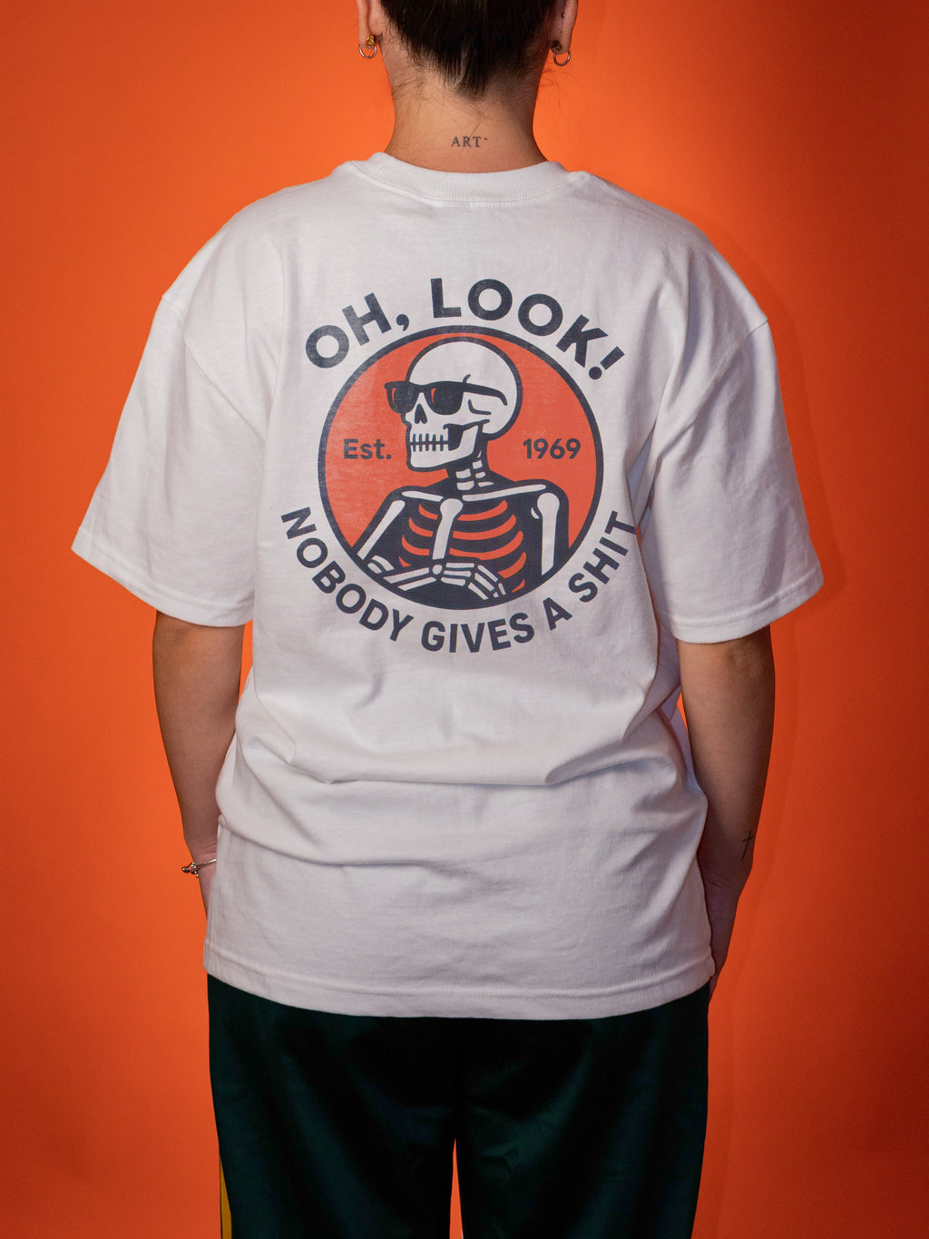 A woman shown from the back wearing a white graphic T-shirt with a large circular skeleton print and text reading 'OH, LOOK! NOBODY GIVES A SHIT' on a solid orange background.