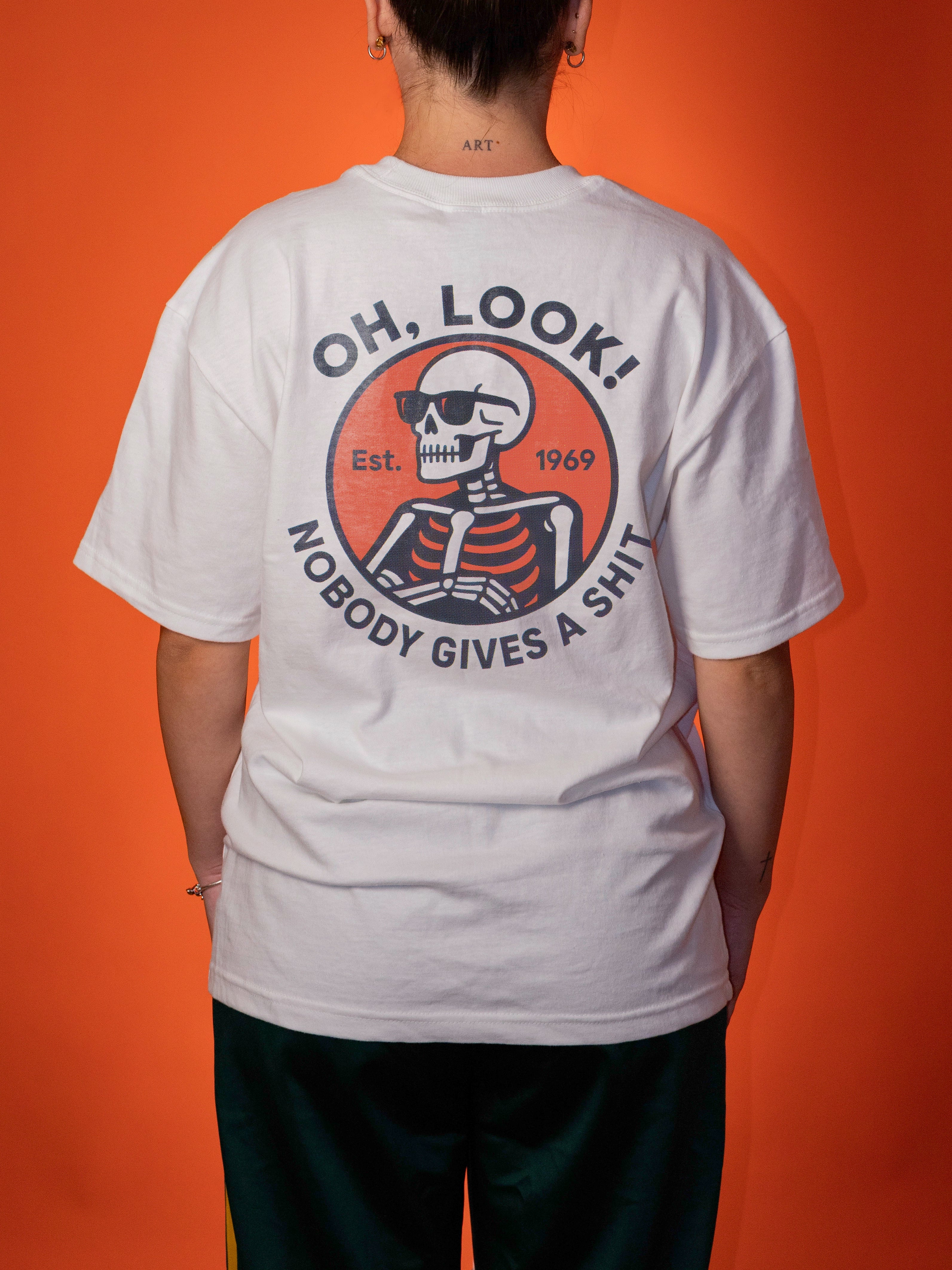 A woman shown from the back wearing a white graphic T-shirt with a large circular skeleton print and text reading 'OH, LOOK! NOBODY GIVES A SHIT' on a solid orange background.