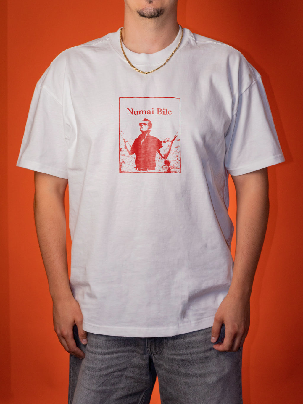 A man wearing an oversized white t-shirt with a red graphic print on the front that reads 'Numai Bile' and features a vintage-style portrait. He is standing against a solid orange background.