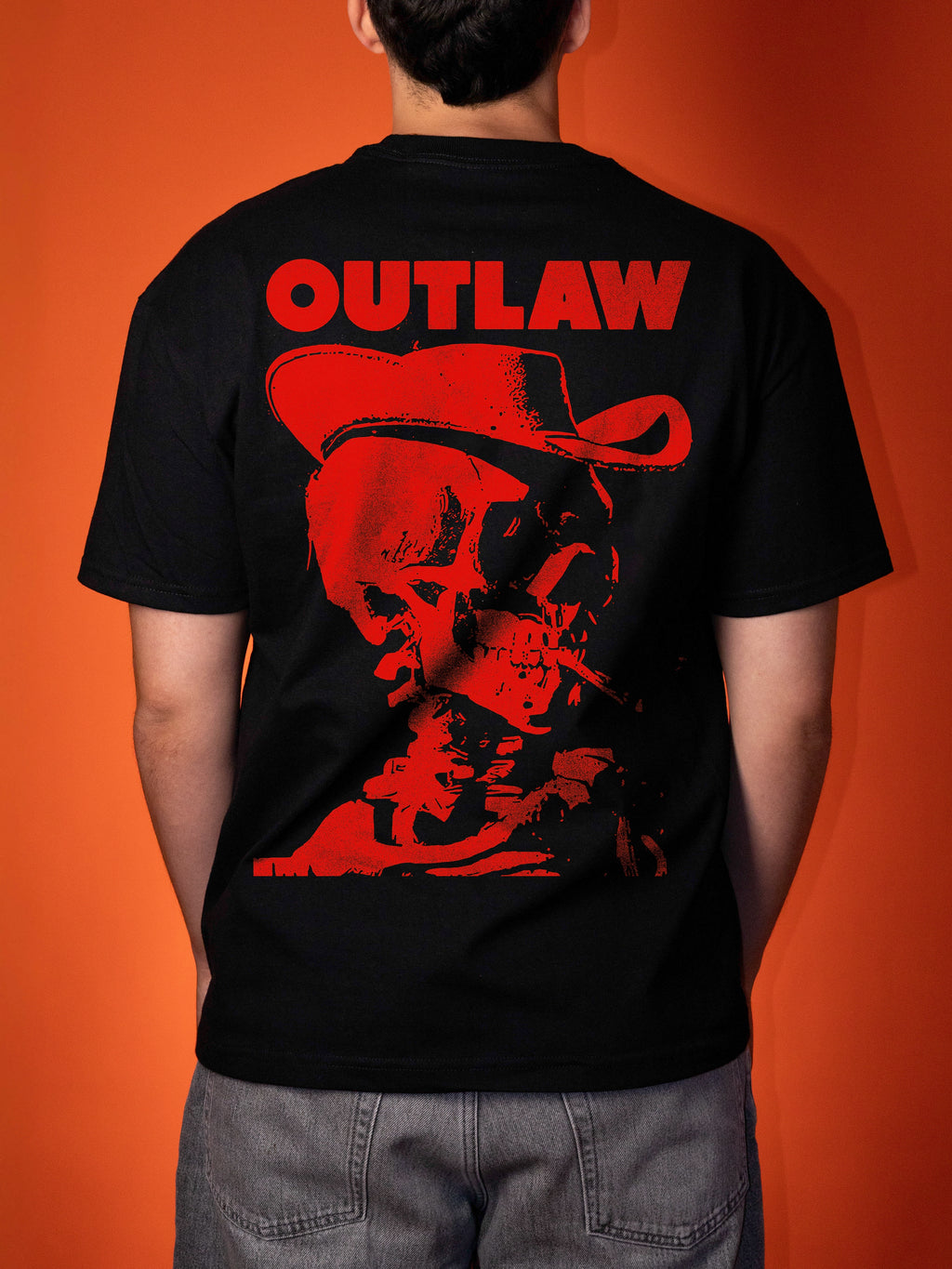 A person viewed from the back wearing a black short-sleeve T-shirt with a large red graphic print on the back featuring the word 'OUTLAW' above a red cowboy hat and skull illustration, against an orange background.