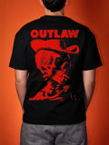 A person viewed from the back wearing a black short-sleeve T-shirt with a large red graphic print on the back featuring the word 'OUTLAW' above a red cowboy hat and skull illustration, against an orange background.