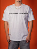 A man wearing a white short-sleeve t-shirt with the text 'I'M SCARED OF WOMEN' printed in black across the chest, paired with gray jeans and a gold chain necklace, set against a solid orange background.