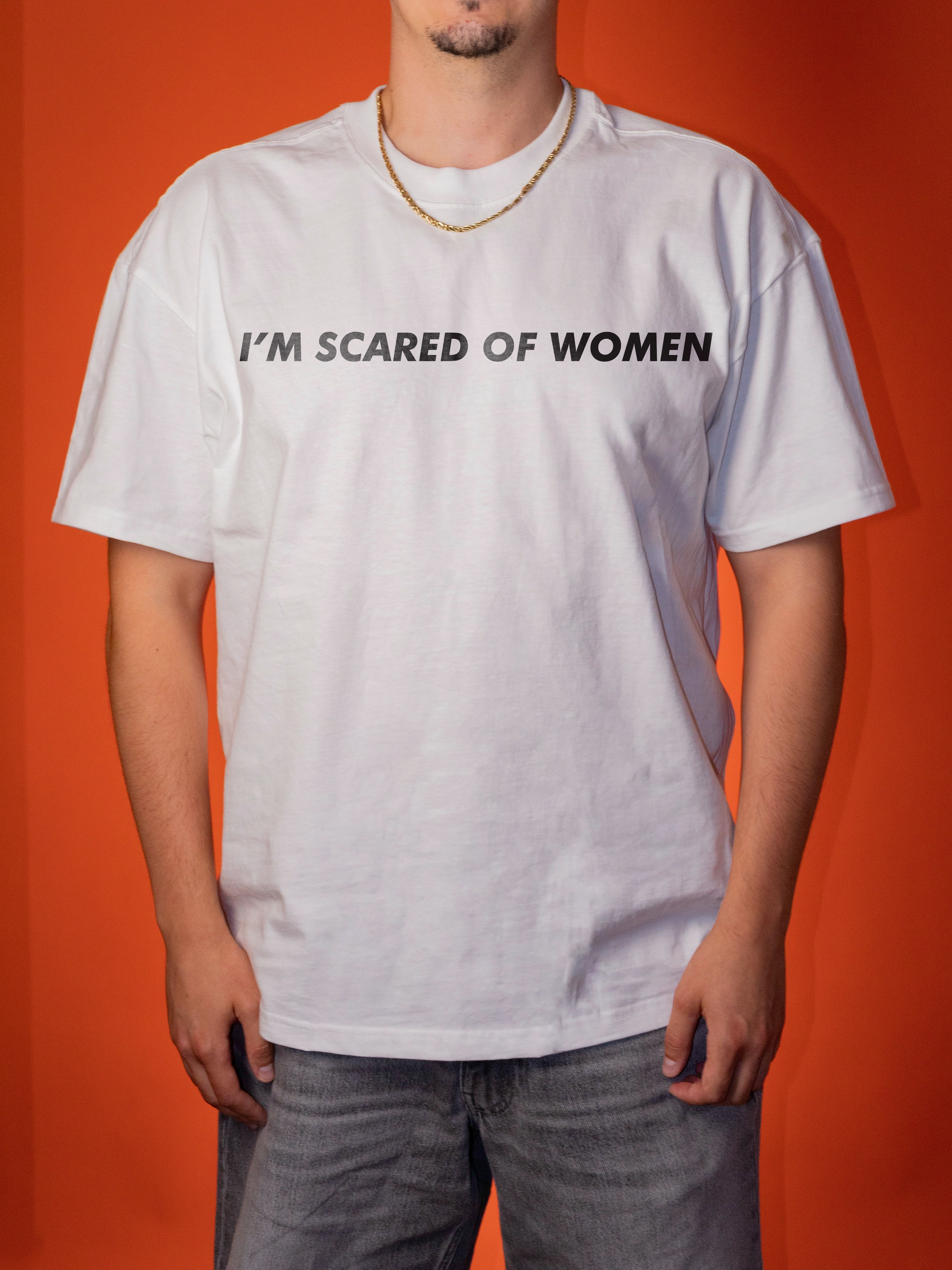 A man wearing a white short-sleeve t-shirt with the text 'I'M SCARED OF WOMEN' printed in black across the chest, paired with gray jeans and a gold chain necklace, set against a solid orange background.