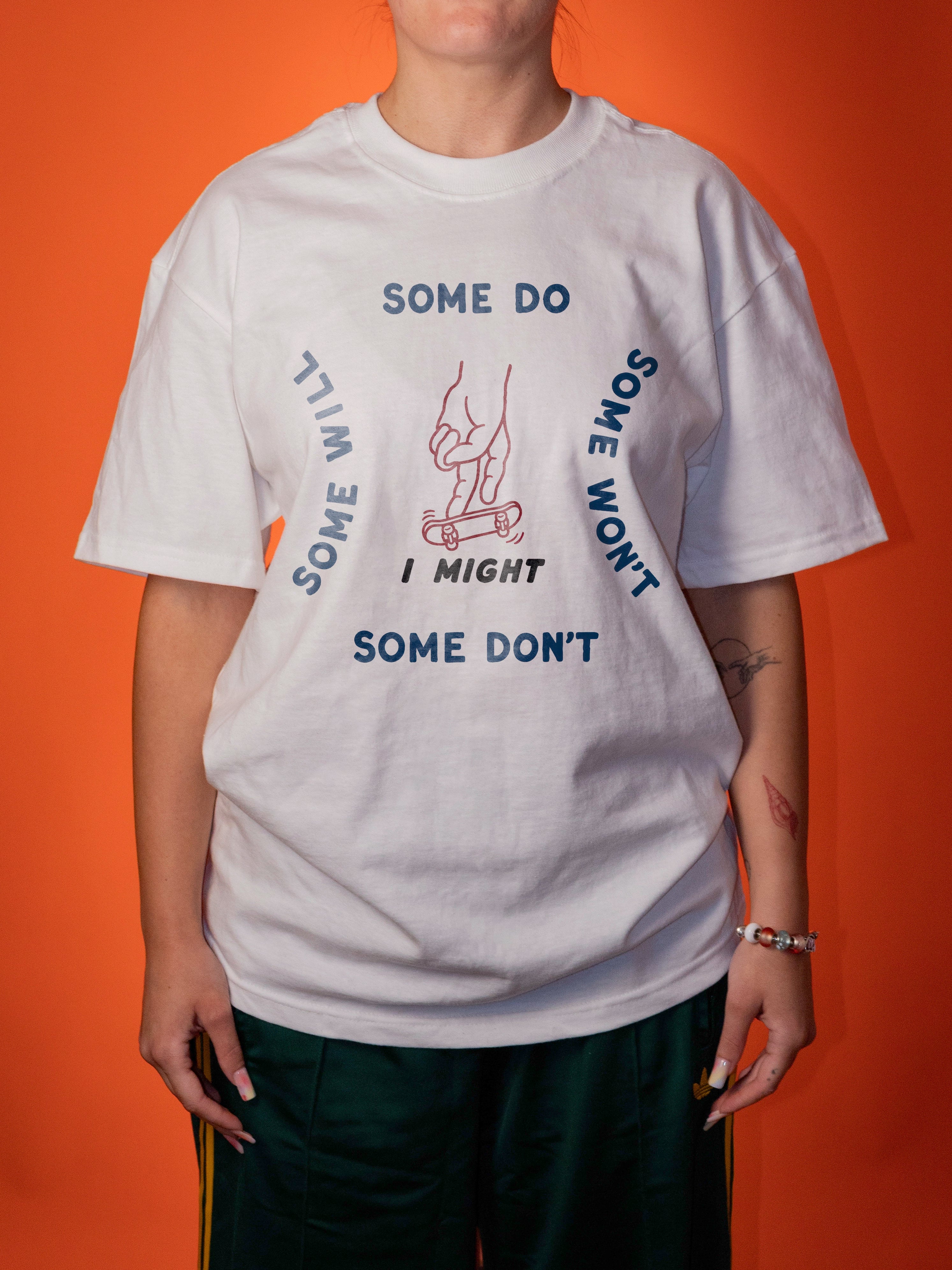 A person wearing an oversized white graphic T-shirt with blue text reading 'SOME DO SOME WON'T SOME WILL SOME I MIGHT I MIGHT' around a hand holding a skateboard, paired with dark green pants, against a solid orange background.
