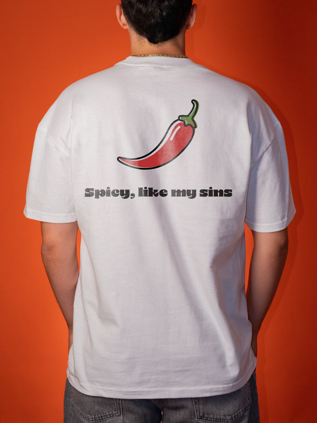 A man viewed from behind wearing a white t-shirt with a graphic of a red chili pepper and the text 'Spicy, like my sins' printed on the back, set against a solid orange background.