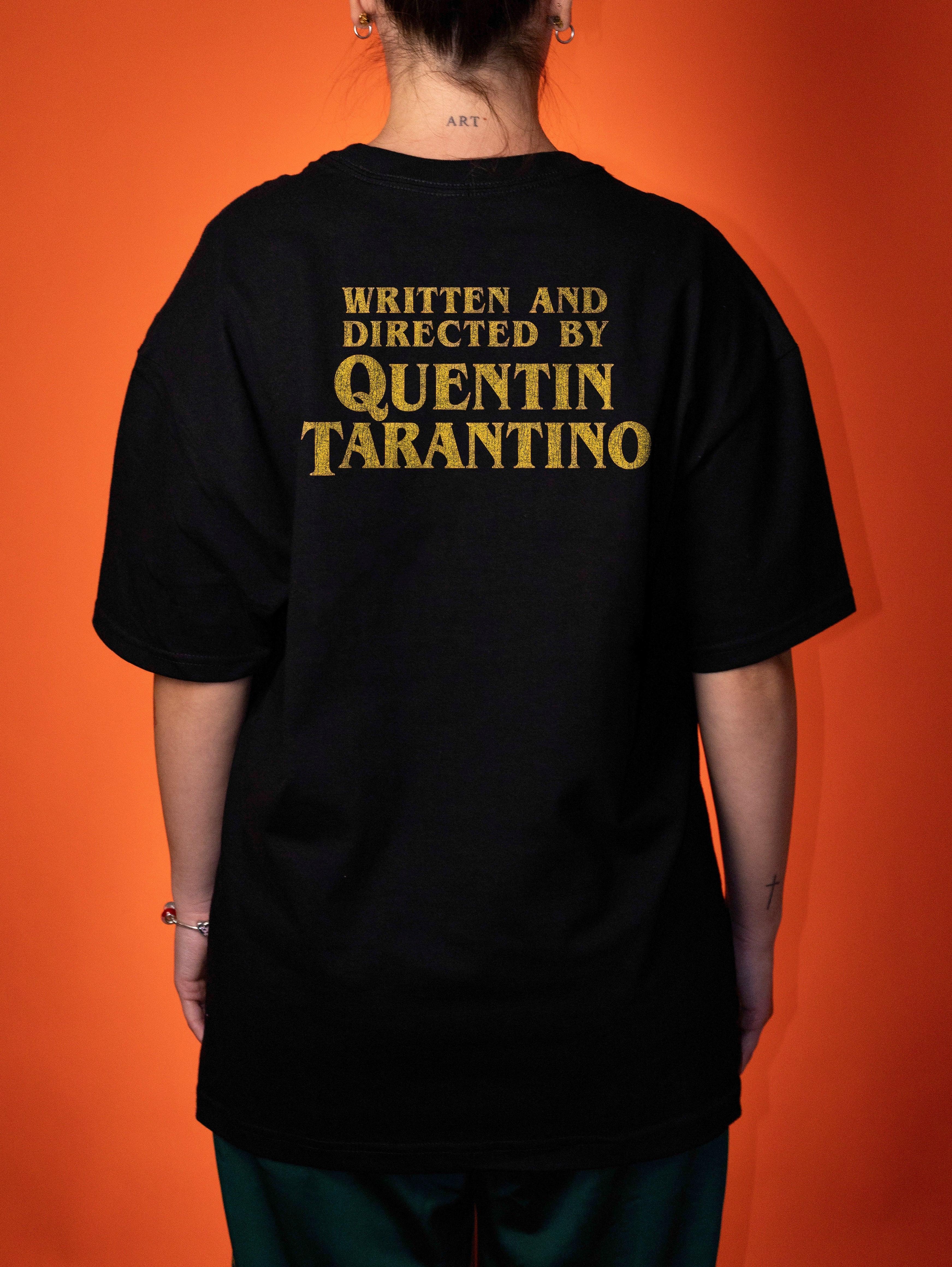 A person shown from the back wearing a black oversized T-shirt with distressed gold text reading 'WRITTEN AND DIRECTED BY QUENTIN TARANTINO' against a solid orange background.