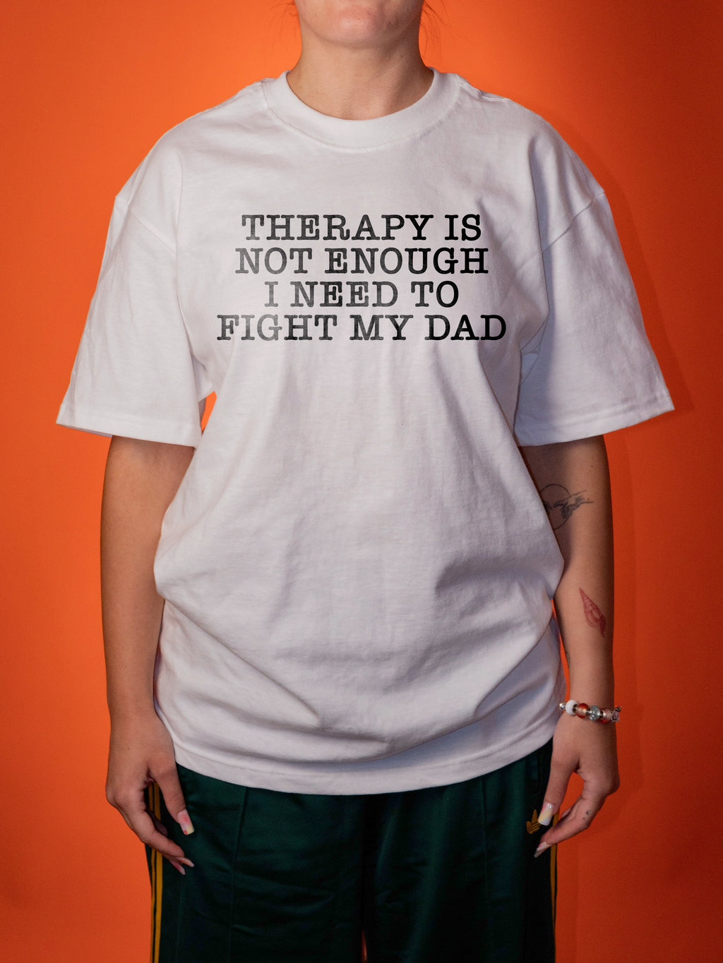 A person wearing an oversized white graphic T-shirt with black text that reads 'THERAPY IS NOT ENOUGH I NEED TO FIGHT MY DAD', paired with dark green pants, standing against a solid orange background.