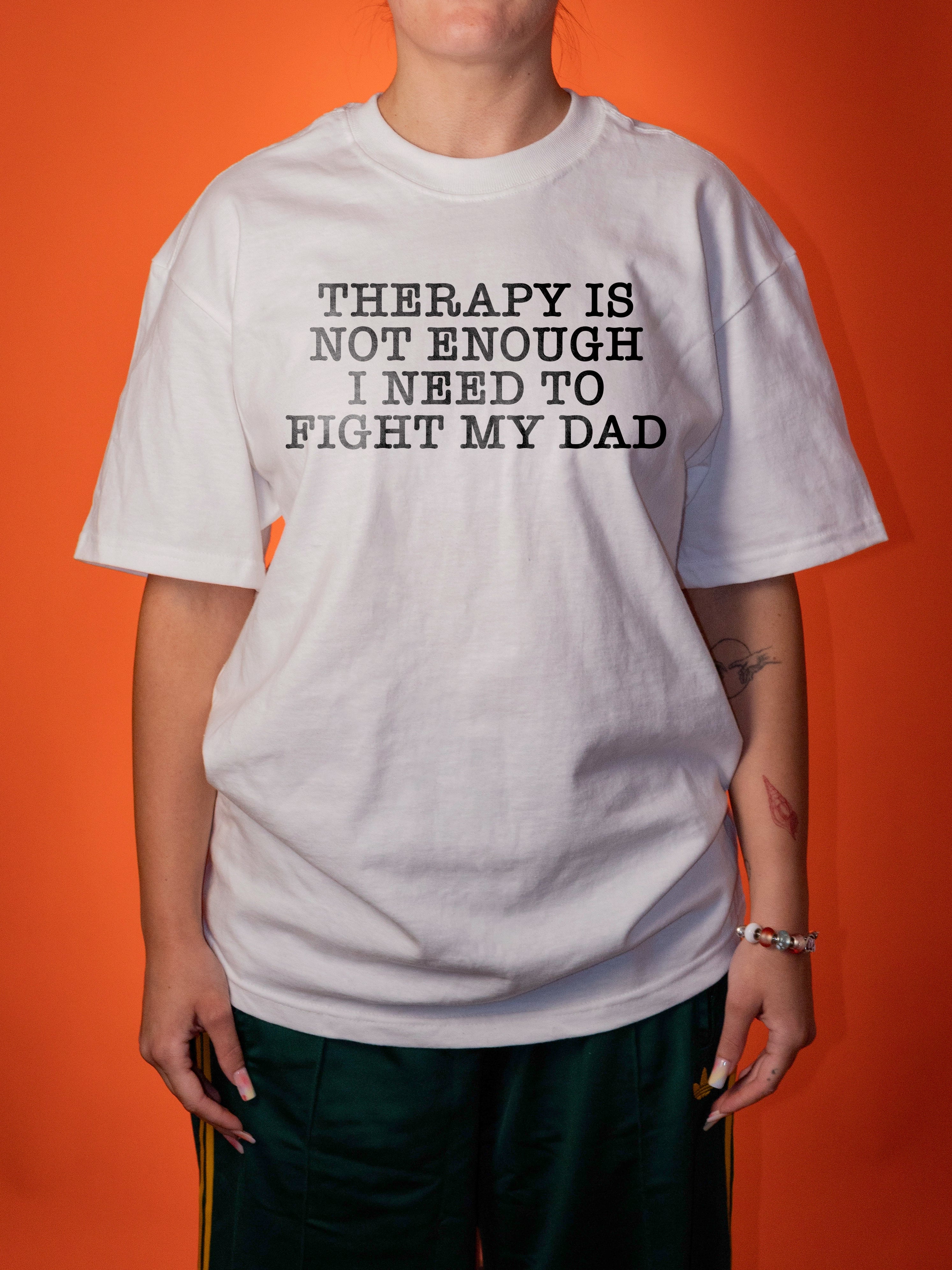 A person wearing an oversized white graphic T-shirt with black text that reads 'THERAPY IS NOT ENOUGH I NEED TO FIGHT MY DAD', paired with dark green pants, standing against a solid orange background.