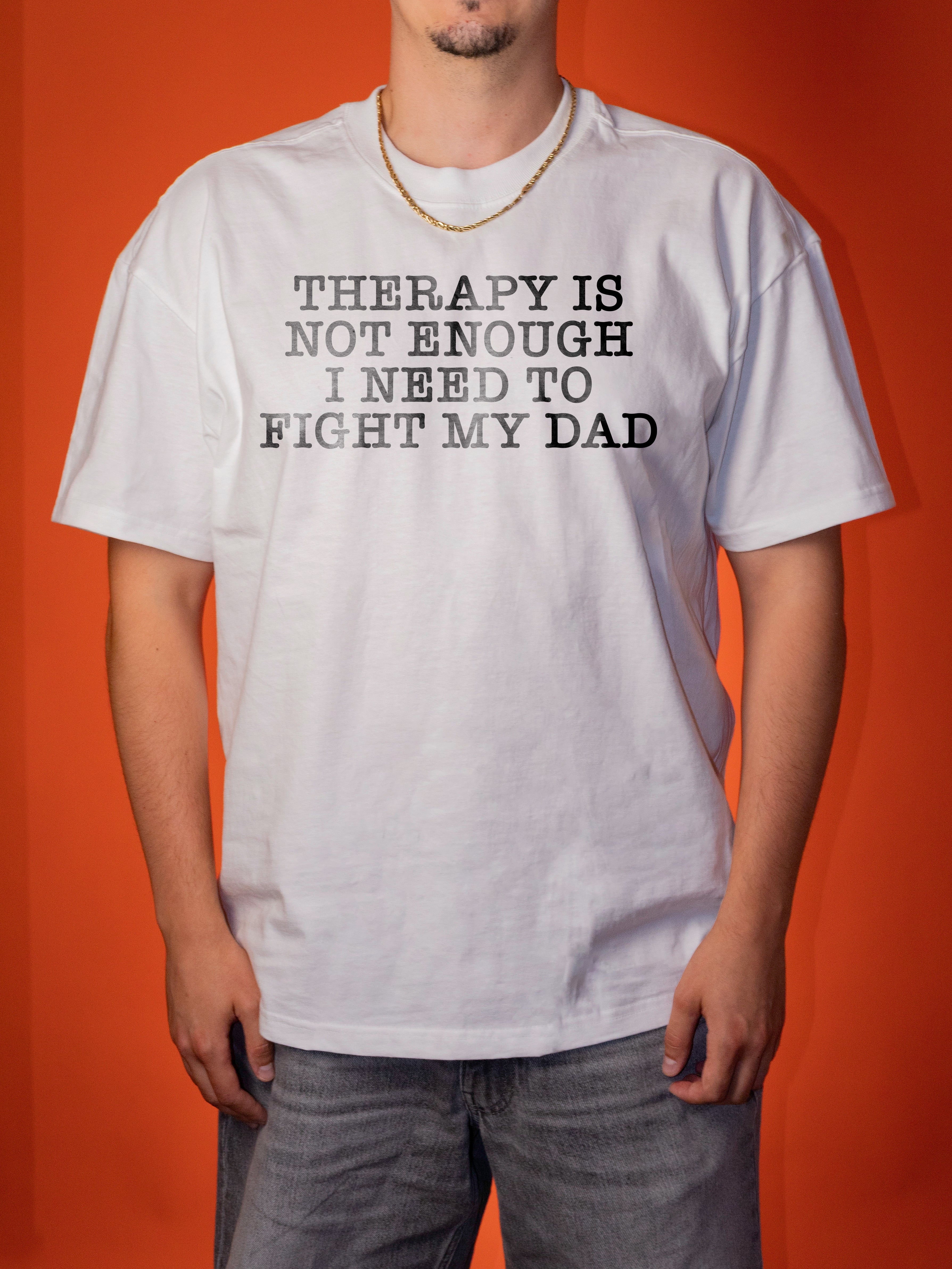 A man wearing a white graphic t-shirt with the text 'THERAPY IS NOT ENOUGH I NEED TO FIGHT MY DAD' printed in black distressed lettering, set against a solid orange background.