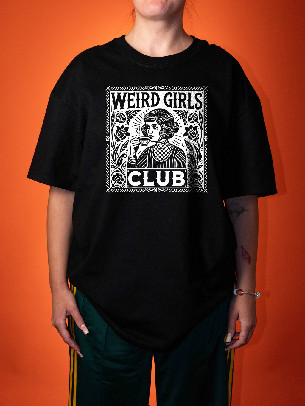 A person wearing an oversized black graphic T-shirt with a white vintage-style illustration and the text 'WEIRD GIRLS CLUB' on the front, paired with dark green pants, against a solid orange background.