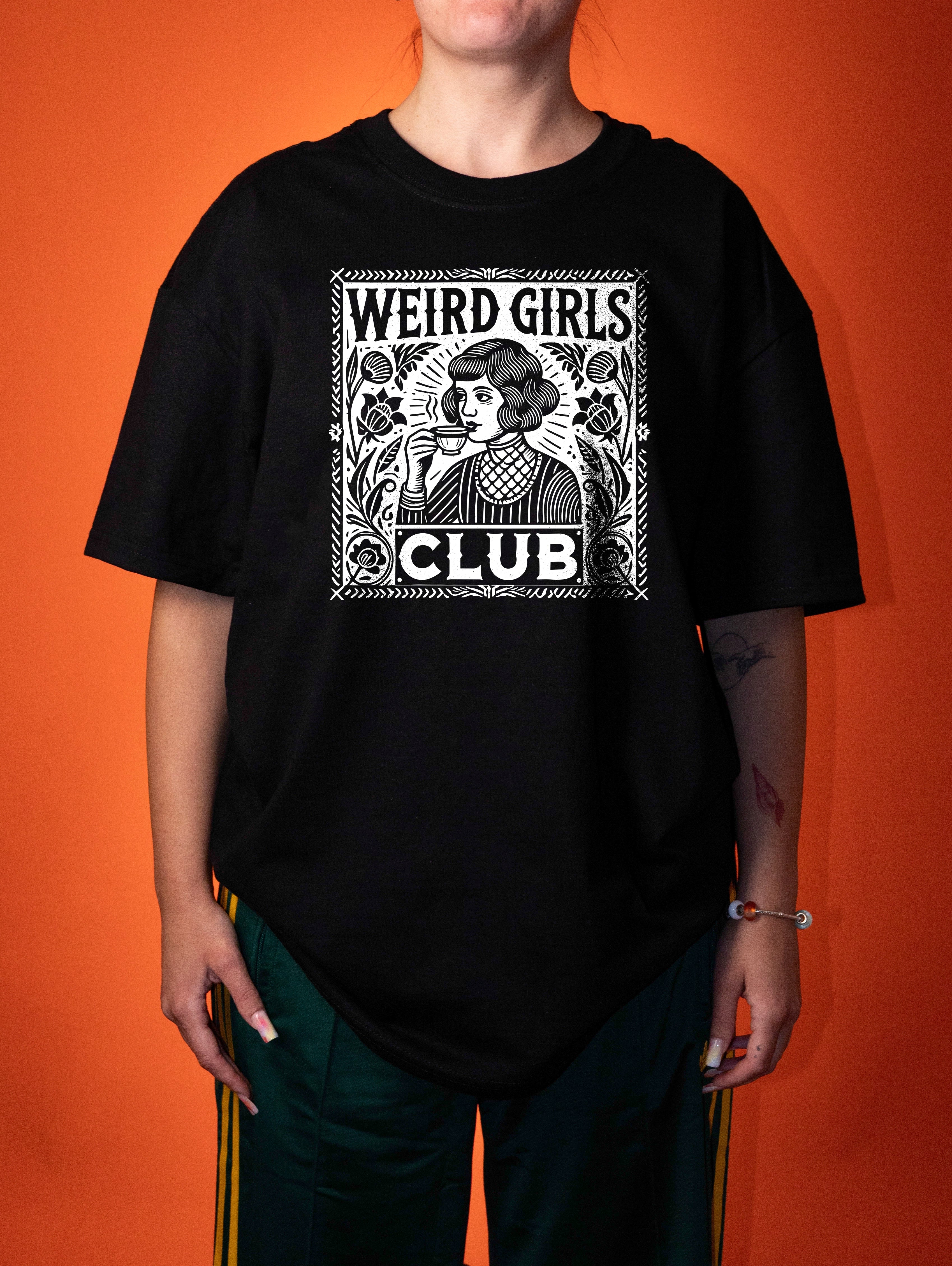 A person wearing an oversized black graphic T-shirt with a white vintage-style illustration and the text 'WEIRD GIRLS CLUB' on the front, paired with dark green pants, against a solid orange background.