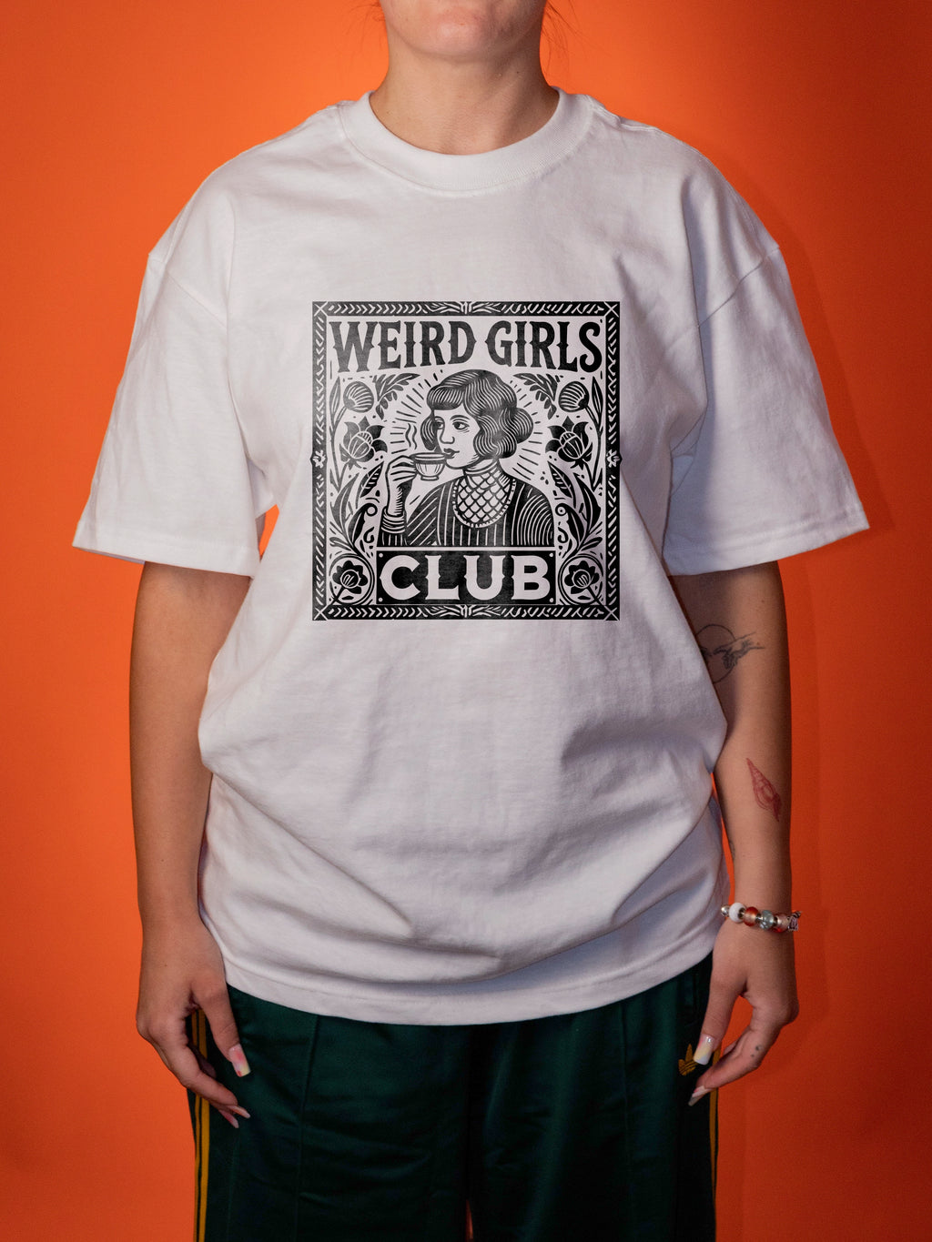 A person wearing an oversized white graphic T-shirt with a black vintage-style illustration and the text 'WEIRD GIRLS CLUB' on the front, paired with dark green pants, against a solid orange background.