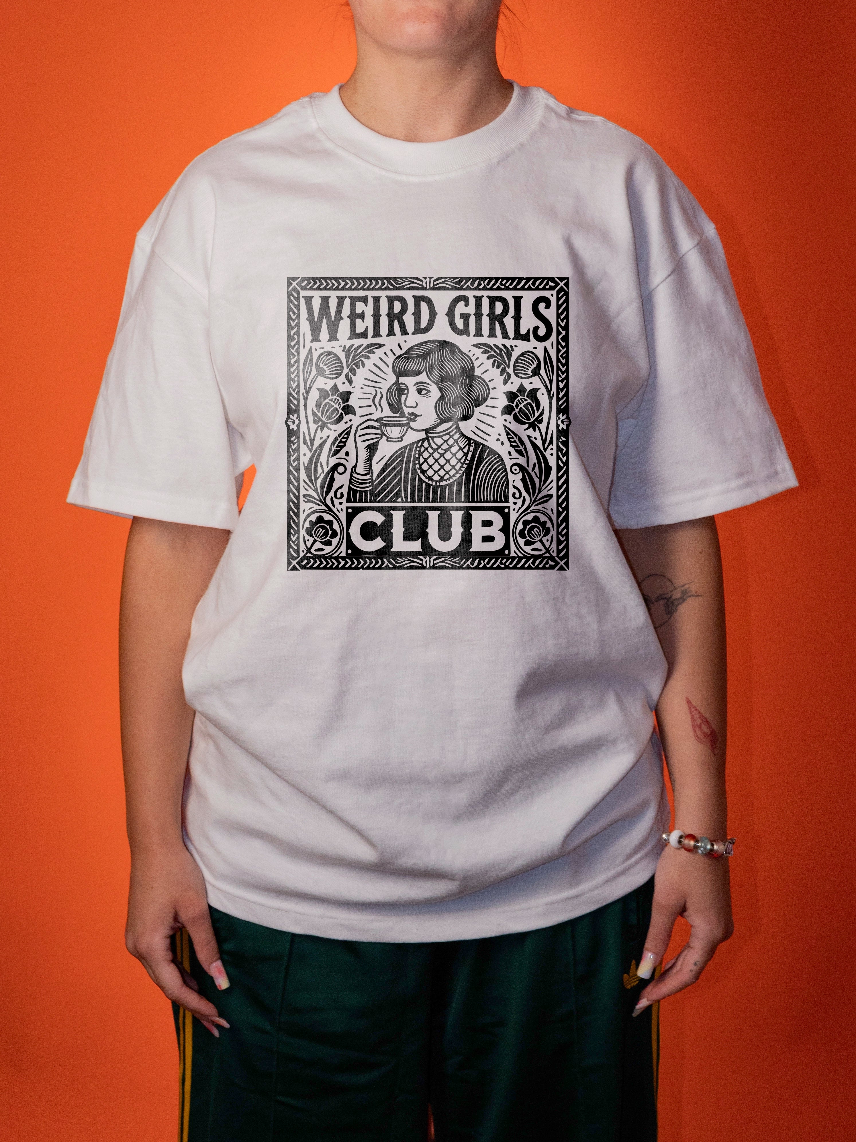 A person wearing an oversized white graphic T-shirt with a black vintage-style illustration and the text 'WEIRD GIRLS CLUB' on the front, paired with dark green pants, against a solid orange background.