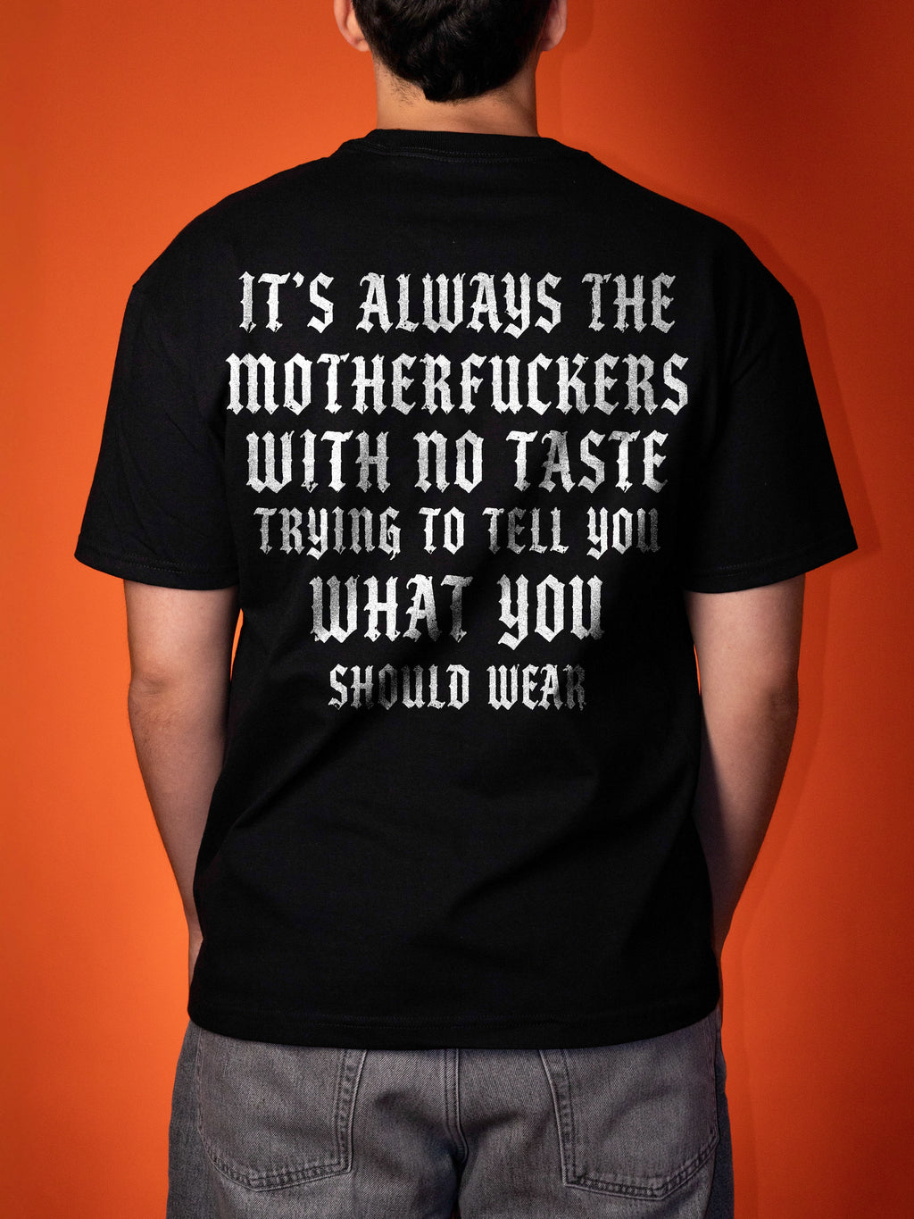 A person shown from the back wearing a black graphic t-shirt with a large white gothic-style text print on the back, set against a solid orange background.