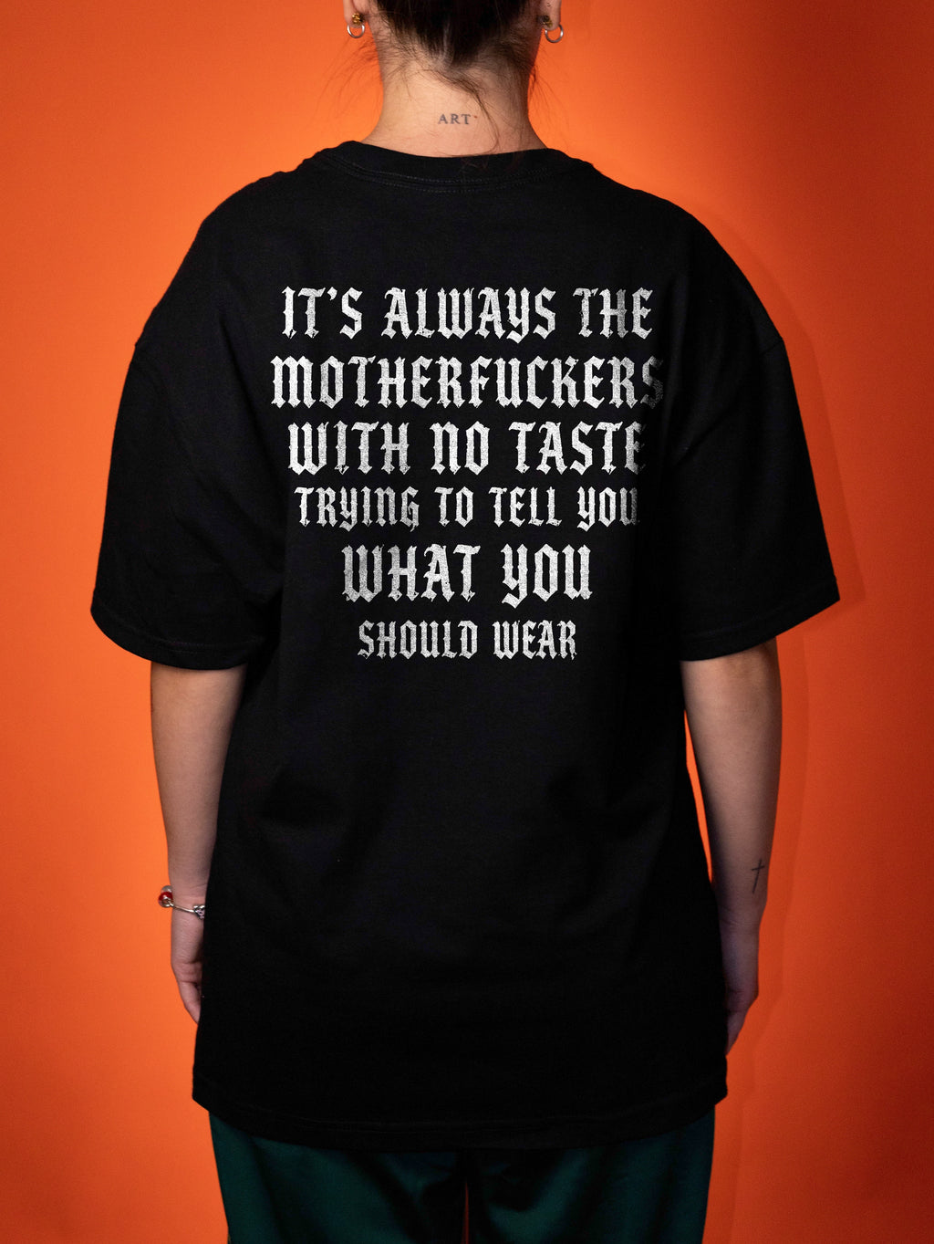A person viewed from the back wearing an oversized black T-shirt with large white text on the back, paired with dark green pants, set against a solid orange background.