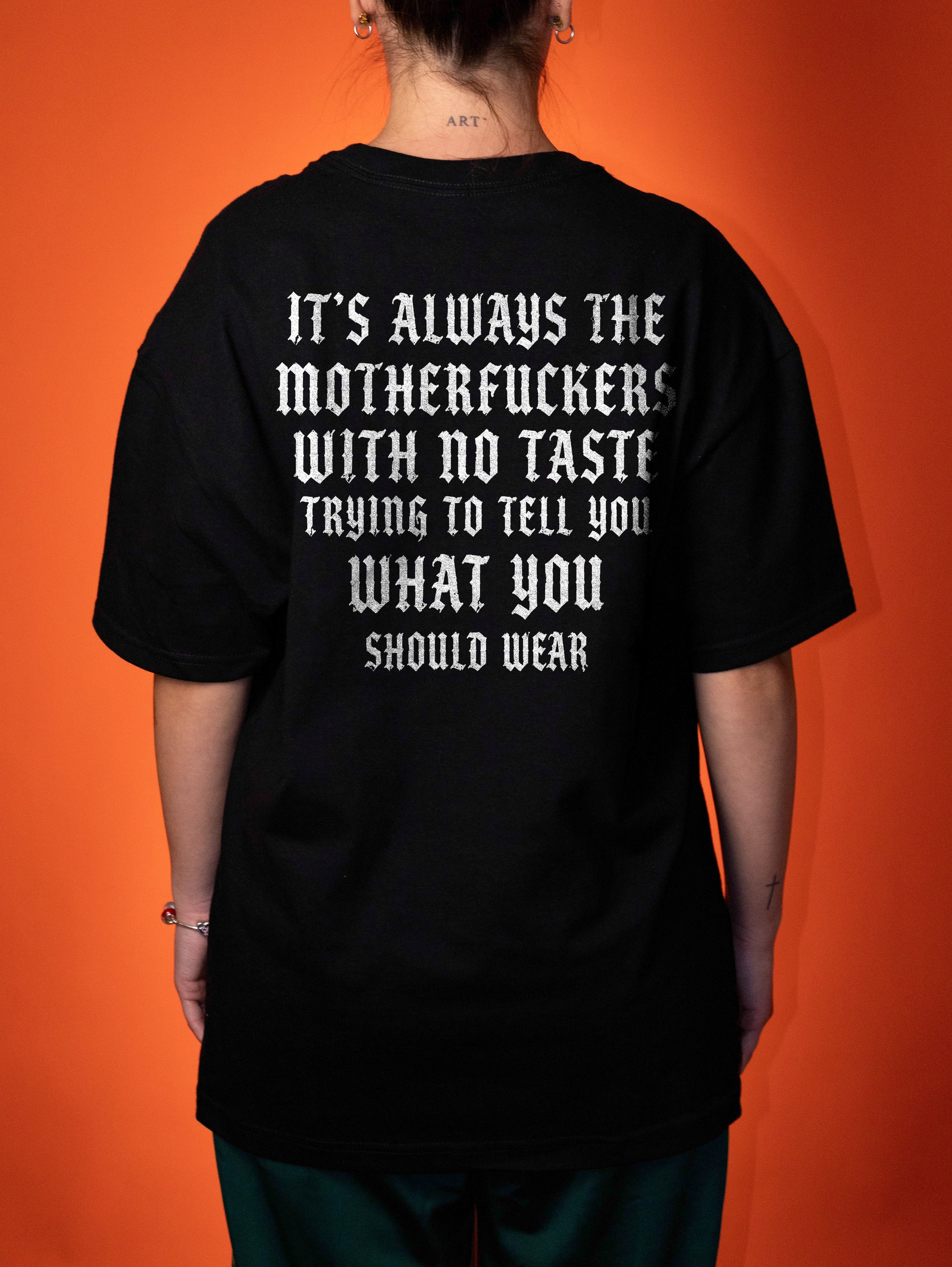 A person viewed from the back wearing an oversized black T-shirt with large white text on the back, paired with dark green pants, set against a solid orange background.