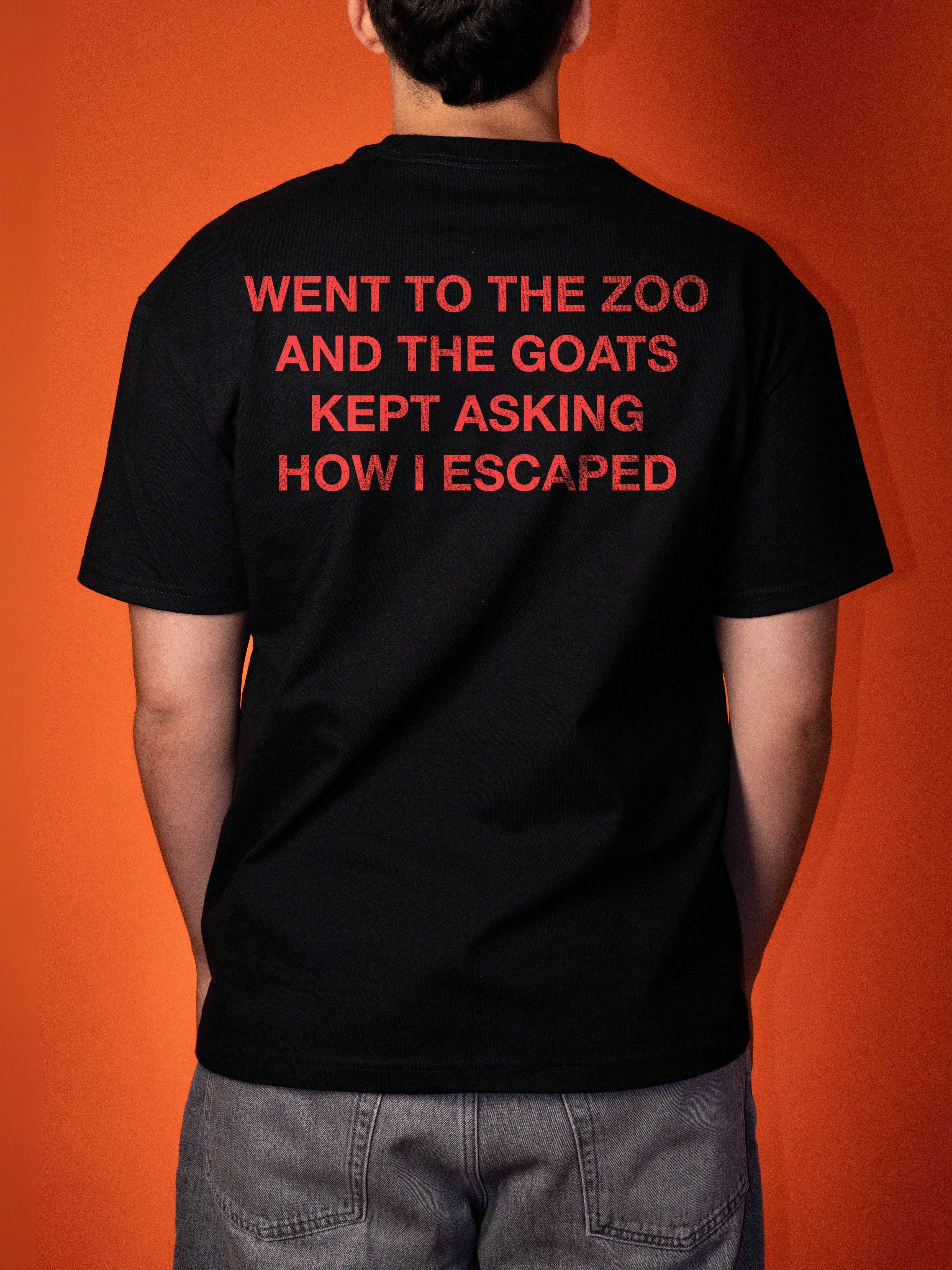 A person shown from the back wearing a black t-shirt with red text that reads 'WENT TO THE ZOO AND THE GOATS KEPT ASKING HOW I ESCAPED' against a solid orange background.