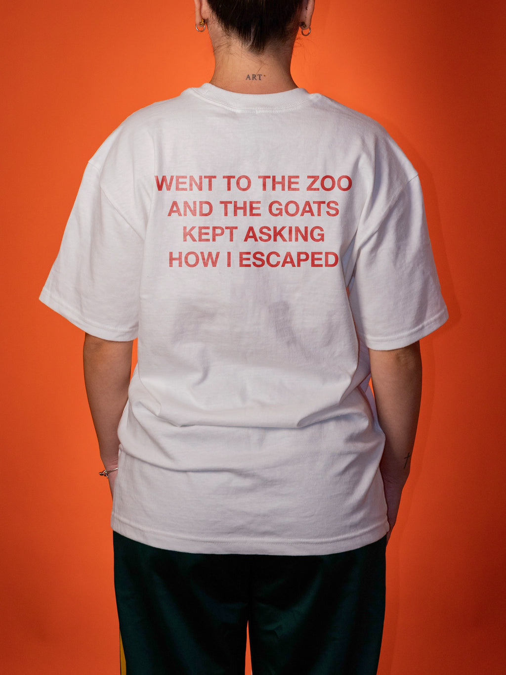 A person shown from the back wearing a white t-shirt with red text that reads 'WENT TO THE ZOO AND THE GOATS KEPT ASKING HOW I ESCAPED' against a solid orange background.